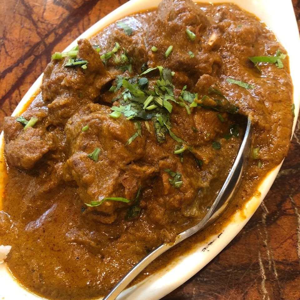 Chettinadu Indian Cuisine's rich brown curry with meat pieces, garnished with fresh cilantro in a white dish.