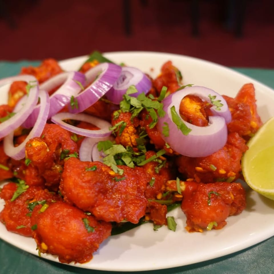 Chettinadu Indian Cuisine's bright red fried chicken, garnished with red onion rings, cilantro, and a lime wedge.